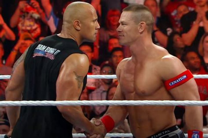 Dwayne Johnson and John Cena wrestle in the ring. Johnson has his hands on Cena's face and Cena has his arms wrapped around Johnson's waist.